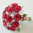 In this image, a classic dome of red roses and white gypsophila is shown as if it has just been placed on a polished table in an elegant Epping home overlooking the edge of Epping Forest. The roses are a deep, romantic crimson, each bloom at that ideal stage where the petals are open enough to reveal their layered structure, yet still tight enough to look plush and full. Their surfaces appear almost satin-like, catching the gentle daylight so that subtle highlights trace each curve. Intertwined between the roses, sprays of white gypsophila-often called baby's breath-form tiny clouds of blossom, each floret no larger than a pinhead, creating a soft, misty frame that contrasts with the bold, confident roses. The greenery is minimal and carefully chosen, small leaves offering a fresh green accent that enhances the red and white without crowding them. The stems are tightly gathered and expertly wrapped, the handle finished with a ribbon coiled neatly and accented with pearls that give a nod to traditional bridal styling. The bouquet feels poised between classic and contemporary, as suitable for a wedding at a local Epping church as for a proposal dinner at a restaurant on Epping High Street. The imagined scent is gentle yet distinctive, a sweet rose aroma that suggests tenderness, commitment, and cherished milestones. Everything about the composition-from the balanced height of the blooms to the precision of the hand-tying-reflects the eye and touch of a florist who understands how to turn fresh flowers into an emotional keepsake.