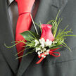 This close-up of a handcrafted wedding boutonnière captures the quiet elegance that a skilled local florist in Epping brings to a special occasion. A hot pink rose forms the central focus, its petals spiralling inward like a soft, protective embrace. The tone is vivid but refined, somewhere between cerise and fuchsia, evoking warmth, affection, and the excitement of a wedding morning. Surrounding the rose are fluffy clusters of white baby's breath, their tiny star-shaped blossoms forming a delicate halo that lightens the design and adds a whisper of countryside charm, like the airy hedgerows you might pass on the lanes approaching Epping Forest. Fine, feathery grasses arc outward in slender loops, giving the arrangement a sense of movement and a modern, slightly whimsical edge. A single deep green leaf, broad and glossy, supports the blooms from behind, its smooth, waxy surface reflecting a gentle sheen that enhances the sense of depth and structure. At the base of the boutonnière, a slim red ribbon is tied in a discreet bow, the satin folds pressed neatly together, suggesting care and intention rather than showiness. The piece rests against a dark, finely woven suit, whose close texture and muted tone make the colours of the flowers appear even fresher and brighter. This boutonnière feels designed for a groom or honoured guest at a wedding near Epping High Street or a reception tucked away in the Essex countryside, quietly stating that every detail-from the first buttonhole to the final toast-has been thoughtfully considered and lovingly prepared.