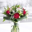 A lush, romantic bouquet stands proudly in a clear cylindrical glass vase, its freshly cut green stems visible through the water like a neat bundle of ribbons. The arrangement is a rich tapestry of deep red and crisp white flowers, glowing softly as though set beside a bright kitchen window in an Epping townhouse. At its heart, velvety red roses unfurl in perfect spirals, their petals layered and generous, radiating that timeless feeling of love and devotion. A large crimson carnation anchors the front, its tightly frilled petals creating a ruffled, almost fabric-like texture that invites the fingertips. Around these passionate reds cluster white alstroemeria, their slender petals subtly speckled inside, alongside closed white lily buds that stand tall and elegant, hinting at the promise of blooms to come. Sprays of delicate white gypsophila float between the larger flowers, like tiny stars scattered across the arrangement, adding softness and a gentle sense of celebration. Encircling the base, broad, glossy dark green leaves are artfully wrapped and curved, forming a polished collar that frames the flowers with structure and grace. Eucalyptus-like foliage and pointed green leaves weave upward, their varied shapes and tones of green adding depth, movement, and a sense of natural abundance, reminiscent of a walk through Epping Forest on a bright afternoon. The bouquet is placed on a clean white surface against a bright, softly blurred background, which lets every colour and texture stand out clearly. The overall mood is sophisticated, warm, and welcoming-perfect for an anniversary dinner in a High Street flat, a birthday surprise sent to a friend near Stonards Hill, or simply to bring a sense of heartfelt affection and fresh beauty into a home or office nearby.