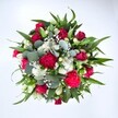 Viewed from directly above against a clean white background, this generous bouquet forms a full, circular burst of colour, as if a garden in Epping High Street had been gathered into a single armful. At the centre, deep crimson red roses curl in layered spirals, some still in bud and others just beginning to open, their velvety petals suggesting quiet romance and thoughtful devotion. Around them, vibrant fuchsia and ruby red carnations with frilled, ruffled edges create a lively texture, each bloom packed with tiny folds that catch the soft, even light. Nestled between the richer tones are clusters of small, daisy-like white spray chrysanthemums, their bright centres and crisp petals lifting the whole design with a fresh, cheerful brightness that feels perfect for a birthday, anniversary, or heartfelt thank you. Threaded delicately through the arrangement are countless tiny white gypsophila flowers, like a gentle mist of baby's breath drifting over the roses and carnations, softening every edge. A frame of lush greenery holds the bouquet together: strong, pointed dark green leaves from lilies or alstroemeria provide structure, while rounded, silvery eucalyptus leaves weave through with a modern, airy touch, their cool blue-green tone evoking a walk through Epping Forest after rain. The overall impression is one of abundance and care, as though an experienced local florist has layered each stem with intention. This is a bouquet that feels both classic and contemporary, ready to be delivered across Epping to celebrate love, lift someone's spirits, or simply bring a vivid splash of nature indoors on an ordinary day.