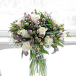 A graceful sympathy bouquet is arranged in a tall, clear glass vase, its straight green stems submerged in clean water and clearly visible, giving the whole design a fresh and uncluttered look. At the centre, several creamy white roses open in soft spirals, their petals smooth and gently curled, conveying tenderness and sincerity. Surrounding these are alstroemeria blooms in soothing shades of purple and lavender, some fully open to reveal softly speckled throats, others in partially closed buds that add variation in height and texture. Fine sprigs of white baby's breath weave throughout, their tiny blossoms forming a light, cloud-like layer that softens and connects each element. The foliage is a rich, natural green, a mix of broad, glossy leaves and more slender stems that cradle the blooms and add depth, echoing the greenery found in the quieter corners of Epping's residential streets. Bathed in bright but gentle natural light, the bouquet stands against a softly diffused white background, as though placed by a window overlooking a calm garden off Station Road. Every petal, leaf, and stem is clearly defined, yet nothing feels harsh or overly dramatic; instead, there is a sense of composure and quiet beauty. This arrangement feels perfectly suited to expressing sympathy, whether delivered to a family home near Epping Forest or placed in a peaceful corner of a room where loved ones gather to remember, bringing a touch of solace and natural grace to a difficult time.