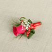 At the centre of this refined boutonniere is a single hot fuchsia rose, captured at just the right moment of bloom, its petals luxuriously full and velvety to the touch. Surrounding it, tiny stars of white baby's breath cluster in soft halos, adding a cloud-like delicacy and gently diffusing the vivid colour of the rose. Scattered among the gypsophila are rich red hypericum berries, smooth and rounded, their glossy skins catching the light and introducing a joyful, almost festive accent. Encircling the flowers, broad loops of deep green foliage and a few slender, arching blades of grass create a sense of graceful movement, as if the boutonniere were shaped by a gentle breeze drifting across the open spaces near Epping Forest. The stems at the base are carefully wrapped in a red ribbon, snug and tidy, with a slight sheen that suggests a touch of ceremony and care. Set against a neutral, softly textured fabric, every element is easy to distinguish: the rose's ruffled edges, the fine stems of gypsophila, the compact berries, the polished greenery. The overall effect is elegant yet approachable, like something chosen for a relaxed wedding reception in a local Epping venue or a special prom evening after photos taken along Epping High Street. This is the sort of buttonhole a local florist would pin with steady hands and quiet pride, ensuring that its colours remain bright and its form intact throughout a day of celebration, photos, and heartfelt embraces.