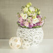 On a smooth, white surface reminiscent of a freshly prepared console in an Epping townhouse, a refined floral arrangement commands attention with its soft, romantic palette. At the centre, large lisianthus blooms unfold in shades of clean white and buttery cream, their multi-layered petals creating a sense of depth and movement, like small clouds captured in bloom. Nestled among them are tall stems of blush pink stock, the clustered florets adding a sense of fullness and quiet fragrance, while small spherical allium blossoms in gentle purple spike out like tiny fireworks, introducing a modern edge. Lush greenery threads throughout the bouquet, with a darker band of leaves carefully wrapped around the base of the flowers, anchoring the design into a rounded, well-balanced dome. The flowers are cradled in an artful spherical vase crafted from slim, white-painted twigs, woven into an open lattice that feels both sculptural and light. Through its airy framework, you can glimpse the stems inside, a detail that hints at the handiwork of a skilled florist familiar with the tastes of local Epping clients. To the left of the vase sit two natural wicker balls in pale wood tones, their crisscross patterns echoing the vase's design and adding a subtle, rustic warmth. The background is a softly blurred, neutral wall-perhaps similar to the calm interiors of homes near Bell Common or Ivy Chimneys-its linen-like texture providing a gentle contrast to the smooth tabletop. The entire composition exudes a quiet sophistication suited to thoughtful gifts, spring celebrations, or a serene accent in a living room where natural light filters in from a garden facing Epping Forest.