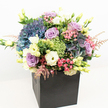 A vibrant, garden-inspired bouquet captures the essence of Epping's lush parks and welcoming spirit, overflowing from a chic matte black square box. The deep indigo and periwinkle hydrangeas, plush and cloud-like, create a dramatic centrepiece, their colours whispering of twilight strolls through Epping Forest. Among them, lavender and blush roses emerge-petals soft and inviting-bringing gentle hints of romance and celebration. Crisp white and buttery yellow lisianthus blooms provide luminous contrast, their delicate trumpets adding a fresh elegance reminiscent of the wildflowers found along Bell Common. Sprays of feathery pink astilbe and lively clusters of lime-green filler flowers interlace throughout, infusing the bouquet with texture and whimsy. Layers of lush, varied greenery frame the blossoms, lending fullness and the invigorating scent of a summer garden after rain. The sharply modern container enhances the bouquet's contemporary style, making it an ideal feature for Epping homes, offices, or as a distinguished gift. Bright, even lighting highlights the rich tapestry of colours and intricate forms, celebrating the skill and creativity of Epping's local floristry scene. Suitable for articles on luxury flower gifts, unique home decor, or same-day floral delivery in Epping, this arrangement embodies sophistication, warmth, and natural beauty in every thoughtfully placed stem.