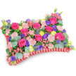 A vividly coloured pillow-shaped floral tribute captured from a slightly elevated, oblique view against a clean white backdrop, the composition crafted to read like a soft, comforting cushion of blooms. The top plane is densely surfaced with velvety hot pink roses-buds and gently unfurling blooms that give a plush, tactile centre-surrounded by clusters of pale, shell-pink carnations whose frilled edges create a delicate, ruffled texture across the face of the cushion. Scattered among them are deep purple lisianthus with their bell-like forms and cheerful lemon-yellow ranunculus that pop like little sunbursts, lending an uplifting warmth to the palette. Cool, architectural notes come from silver-blue eryngium thistles tucked thoughtfully into the mass, their spiky silhouettes bringing contrast and a hint of wildness. Rounds of silvery-green eucalyptus leaves and assorted rich greenery weave through the arrangement, offering a verdant backdrop and a gentle, aromatic lift when passed under the nose. The whole block of flowers sits framed by a pleated, ruffled red satin ribbon base that defines the pillow outline and gives a refined, memorial finish to the piece. Soft, even lighting highlights the sheen of the satin edge and the velveteen petals without harsh shadows, inviting a quiet, consolatory feeling rather than solemn gloom. This traditional funeral pillow reads as a celebration of memory-energetic fuchsia, violet, lemon and blush knit together to convey affection and remembrance-an arrangement a family might choose to honour a loved one at a service, graveside, or placed beside a photograph during a wake in and around Epping.