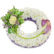 A serene, circular sympathy wreath composed of a dense cloud of small white chrysanthemum florets, tightly packed to create a soft, cushiony surface that reads as calm and pure. Beneath the pristine white a subtle band of deep purple mesh or ruffled netting peeks through along the inner edge, lending quiet depth and a muted, dignified halo. On the upper-left quadrant a compact spray of greenery and flowers is artfully gathered: at its heart a single cream-to-pale-yellow rose unfurls gentle, velvety petals, surrounded by glossy variegated leaves with lighter margins and delicate, feathery fern fronds that add a whisper of movement. Threaded through this cluster are thin sprigs of lavender-hued statice, their tiny clustered blooms contributing a fragile texture and a cool counterpoint to the cream rose. Mirroring the left cluster and balancing the composition, two elegant calla lilies sweep across the right side from about two o'clock to five o'clock; their trumpet-shaped blooms show a graceful gradient from rich, deep purple at the tips to soft peach-pink and blush nearer the bases, while their bright green stems curve along the lower-right arc and tuck neatly into the chrysanthemum base. A single longer green stem, emerging from the left cluster, is secured across the lower wreath and points toward the base of the callas, guiding the eye in a soft, connecting line. Photographed against a clean white background in diffuse daylight, the arrangement reads as a dignified tribute - tactile, refined, and suited to memorial services in Epping, nearby Loughton or Theydon Bois where neighbours often choose classic, understated sympathy pieces.