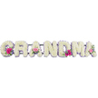 This photograph captures a heartfelt GRANDMA funeral flower tribute created in a classic letter style, where each character is fully covered in pristine white chrysanthemums. The blossoms are arranged so tightly that they form a smooth, cushion-like texture, giving the whole piece a comforting, wrapped-in-love appearance. Around every letter, a delicate lilac ribbon provides a soft outline, lending a refined finish and softly breaking the transition between the white petals and the background. Three focal posies are positioned along the tribute: one at the beginning G, another at a central A, and a final cluster adorning the last A. These petite bouquets are made from bright pink gerbera daisies and tender pink roses, supported by glossy green foliage that adds depth, contrast, and a sense of natural freshness, like the greenery you might see around Epping's open spaces and the edge of the forest. The whole arrangement is set against a plain, light backdrop, with gentle illumination that picks up the slight sheen on the ribbon and the intricate layers of chrysanthemum petals. There is a sense of stillness and reverence in the way the letters are aligned, as though they have just been placed for a service at a local church or crematorium. You can almost imagine the quiet murmur of family and friends remembering shared meals, warm hugs, and stories told over many years. This tribute offers a peaceful and loving way to say farewell to a cherished grandmother, with flowers that echo both purity and tender, enduring love.