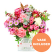 A bright, affectionate floral display fills the frame, as if captured on a clear day in a light-filled Epping living room. At its core, the bouquet features lush pink roses, some in striking magenta and deep fuchsia, others in softer, more romantic blush tones, each bloom showing layers of silky petals that curl and fold like fabric. Ruffled carnations in coral, peach-pink, and pale pastel shades weave around the roses, their frilly edges adding playful texture and a sense of abundance. Smaller daisy-style chrysanthemums in gentle lavender and purple are scattered throughout, their vivid green centres catching the eye and lifting the whole palette with a fresh, almost citrus brightness. Creamy white flowers are interspersed between the colours, providing a soothing contrast that keeps the bouquet harmonious rather than overwhelming. A collar of greenery frames the blossoms: rounded eucalyptus leaves in muted, silvery green sit alongside deeper green foliage and a few reddish-brown sprigs that lend depth and a slightly romantic, evening warmth, reminiscent of the changing light over Epping Forest. All the stems are gathered neatly into a clear, square glass vase, their lines visible through the water. Around the base of the vase runs a white decorative band, printed with tiny red hearts and the phrase love all my love, making the intention of the gift feel unmistakably heartfelt. In the bottom right corner of the image, an orange circular badge with bold white capital letters reads VASE INCLUDED, indicating that the bouquet arrives as shown, ready to place on a table in a home off Bell Common or a cosy flat near Epping station. The overall mood is joyful, tender, and perfectly suited to birthdays, anniversaries, or quiet gestures of appreciation.