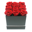 This image presents a striking, modern rose design with a sense of calm precision that would feel at home in many Epping living rooms and townhouses. At the heart of the photograph sits a sleek square gift box finished in a matte black tone, its clean lines giving it the look of a chic hatbox or a small luxury gift. A fine white stripe encircles the lower portion of the box, adding a subtle accent that gently breaks the darkness without distracting from the flowers above. Emerging from the open top are twelve red roses, arranged with almost architectural accuracy in three rows of four. Each bloom is fully unfurled, the petals curled in layered whorls that create a plush, velvety surface. The colour is a rich ruby red, deepening toward the centres where the shadows gather, suggesting preserved or long-lasting roses chosen for their ability to hold both shape and hue over time. The flowers extend just beyond the rim of the box, forming a low, even cushion of colour that invites a second look. Soft, even lighting flows across the composition, picking out the gentle curves of the petals and the subtle texture of the matte box, while the pure white background keeps every detail crystal clear. The result is a mood of understated luxury and deep affection, perfect for an anniversary dinner in a home off Bower Hill, a romantic surprise sent to Epping Upland, or a refined thank-you gift delivered across the CM16 area. The arrangement feels deliberate, thoughtful, and enduring, echoing the kind of meaningful gestures that local residents often entrust to their neighbourhood florists.